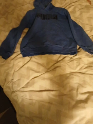 Blå hoodie från Puma - Säljer en blå hoodie från Puma med svart logga på bröstet. Hoodien har en klassisk känguruficka framtill och en skön huva. Perfekt för dig som gillar sportig och avslappnad stil.