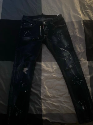 Mörkblå slim fit jeans med slitningar - Snygga mörkblå slim fit jeans med slitningar och vita färgstänk. Jeansen har dragkedja fram, klassiska fem fickor och en cool, lite sliten look. Perfekta för dig som gillar streetstyle och vill sticka ut lite extra.