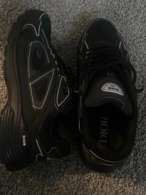 Svarta Dior sneakers med vita detaljer - Säljer ett par svarta Dior sneakers med vita kontrastdetaljer och logga på sidan och plösen. Skorna har en chunky sula och är tillverkade i en mix av mesh och läder för en modern look. Perfekta för dig som vill ha något exklusivt och trendigt.