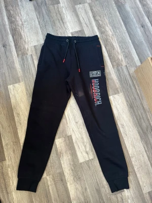 Svarta Hoodrich mjukisbyxor - Säljer ett par svarta mjukisbyxor från Hoodrich med tryck i vitt och rött på ena benet. Byxorna har resår i midjan med dragsko, muddar vid bensluten och sidofickor. Perfekta för en avslappnad streetwear-look.
