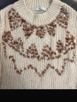 Beige stickad tröja med paljetter Zara - Beige stickad tröja från Zara med rund hals och dekorativa bronsfärgade paljetter och broderier i ett mönster över bröstet och ärmarna. Tröjan har grovstickad struktur och ribbade muddar vid ärmslut och nederkant. Perfekt för dig som gillar detaljer!