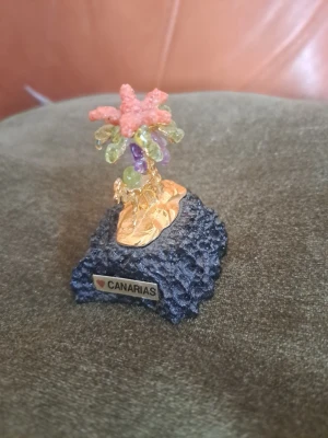 Vackert hantverk från Kanarieöarna – Ädelstensträd på Lavasten - Säljer ett klassiskt "lyckoträd" inköpt på Kanarieöarna. Ett fint prydnadsföremål som kombinerar öarnas vulkaniska natur med vackra mineraler.  Blad: Äkta halvädelstenar i form av grön Olivin (Peridot) och lila Ametist. Toppen pryds av en rosafärgad korall-liknande sten.  Stam: Guldfärgad metalltråd som går att böja för att forma trädet efter egen smak.  Bas: Äkta svart, porös lavasten med texten "Canarias".