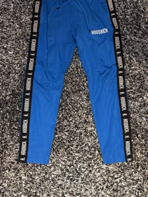 Blå mjukisbyxor från Hoodrich - ⚠️dom är lite sönder över fickan som sitter inne på benet men det syns inte. Där e också lite vit färg på högra benet de syns lite men de är inget man tänker på om man inte säger de⚠️ Säljer ett par blå mjukisbyxor från Hoodrich med svarta sidor där det står HOODRICH i vitt. Byxorna har dragsko i midjan och är tillverkade i ett mjukt och stretchigt material. Perfekta för en chill och avslappnad stil.