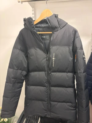 Sail Racing vinterjacka  - Snygg svart dunjacka från Sail Racing med huva och dragkedja framtill. Jackan används inte och är i mycket bra skick.