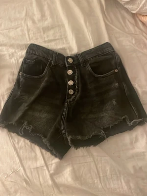 Svarta högmidjade jeansshorts - Säljer ett par svarta högmidjade jeansshorts med fransig kant och slitna detaljer. Shortsen har fem knappar framtill, klassiska fickor både fram och bak samt en cool vintage look. Perfekta för sommaren och riktigt snygga till sneakers eller boots.