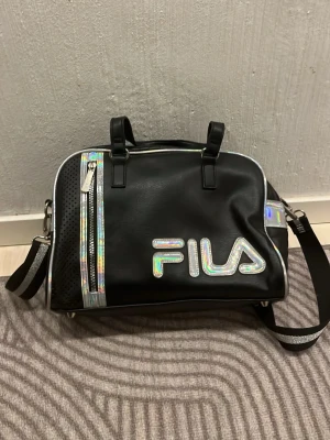 Svart handväska från FILA med holografiska detaljer - Cool svart handväska från FILA med holografiska detaljer och stort logotryck på framsidan. Väskan har både handtag och justerbar axelrem, samt flera fack med dragkedja. Materialet är konstläder och detaljerna ger en futuristisk vibe.