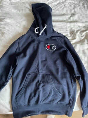 Mörkblå hoodie från Champion - Snygg mörkblå hoodie från Champion med stor logga på bröstet och vit dragsko i huvan. Klassisk känguruficka framtill och mjukt material som känns skönt mot huden. Perfekt för en avslappnad och sportig stil.