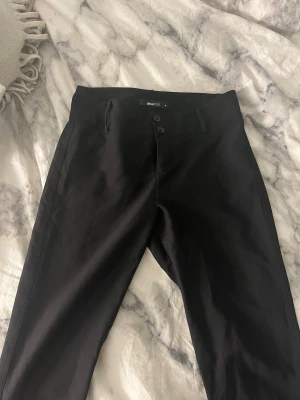 Svarta kostymbyxor från Gina Tricot  - Snygga svarta kostymbyxor från Gina tricot  med utsvängda ben och låg midja. Byxorna har två knappar i midjan och diskreta fickor på sidorna. Materialet är slätt och ger en stilren look. Perfekta för dig som vill ha en clean och modern stil.