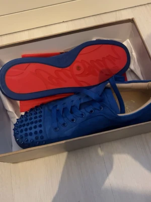 Blåa Louboutin sneakers med nitar - Säljer ett par blåa sneakers från Christian Louboutin med coola nitar på tån och klassisk röd sula. Skorna är låga och har en unik design med matchande blå skosnören och ovandel i mocka. Perfekta för dig som vill sticka ut.