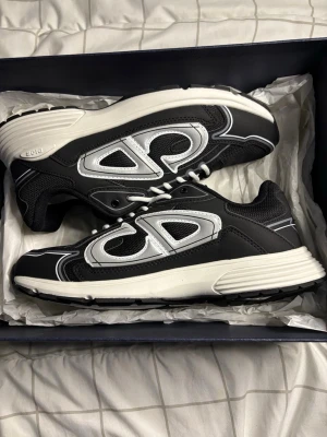 Svarta Dior sneakers med silverdetaljer - Snygga svarta sneakers från Dior med vita och silvriga detaljer samt CD-logga på hälen. Skorna har en chunky vit sula och ovandel i mesh och läder för en modern look. Perfekta för dig som vill sticka ut med exklusiva och trendiga sneakers.