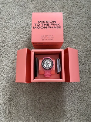 Omega x Swatch Pink Moonphase klocka - Säljer en Omega x Swatch MoonSwatch Mission to the Pink Moonphase. Klockan har rosa boett och armband, vit urtavla med svarta och rosa detaljer samt svart bezel. Tillverkad i biokeramik och levereras i originalförpackning. Perfekt för dig som gillar unika accessoarer.