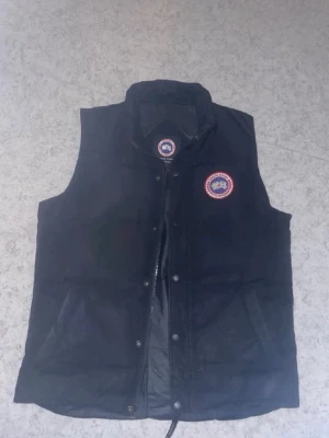 Svart dunväst från Canada Goose - Snygg svart dunväst från Canada Goose med klassisk logga på bröstet. Västen har hög krage, dragkedja och tryckknappar framtill samt två fickor. Perfekt för lager-på-lager och håller dig varm under kyliga dagar.