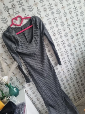 Grå ribbad långklänning - Snygg grå långklänning med ribbad struktur och djup v-ringning. Klänningen har långa ärmar och är figurnära i modellen, perfekt för dig som gillar en stilren och trendig look.