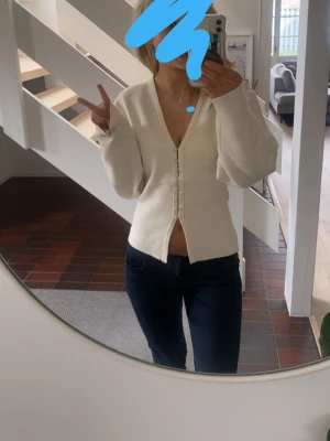 Vit kofta med knappar och v-ringning - Säljer en stilren vit kofta med djup v-ringning och dekorativa knappar framtill. Koftan har långa ärmar och en något croppad passform som ger en trendig siluett. Perfekt att matcha med jeans eller kjol för en clean look.