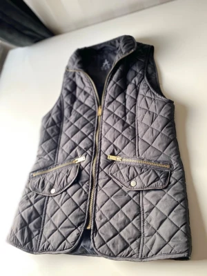 Svart quiltad dunväst med dragkedja - Stilren svart dunväst med quiltat mönster och hög krage. Västen har två snedställda fickor med dragkedja och tryckknapp, samt guldfärgade detaljer. Perfekt att slänga över en hoodie eller tröja för extra värme.