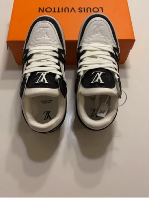 Louis Vuitton svartvita sneakers - Säljer ett par Louis Vuitton sneakers i svart och vitt med tydliga LV-loggor på tungan och sidan. Skorna har klassisk låg profil, vita snören och detaljer i svart läder med monogrammönster. Perfekt för dig som vill sticka ut med exklusiva sneakers.