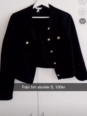 Svart kavaj med guldknappar från H&M - Stilren svart kavaj från H&M med dubbelknäppning och dekorativa guldknappar framtill. Kortare modell med klassisk krage och långa ärmar. Perfekt för att lyfta en outfit med en elegant touch.