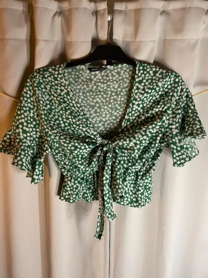 Grön blommig omlottopp med volang - Söt grön omlottopp med vitt blommönster och volangdetaljer. Toppen har korta ärmar, knyt framtill och en luftig, croppad passform. Perfekt för varma dagar och ger en fräsch look till din outfit.