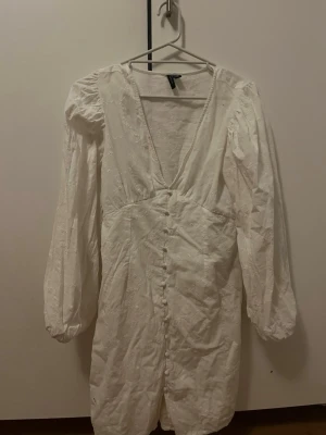 Vit långärmad kortklänning med knappar - Söt vit kortklänning med långa puffärmar och djup v-ringning. Klänningen har dekorativa knappar framtill och ett subtilt broderat mönster i tyget. Perfekt för sommarens alla tillfällen.