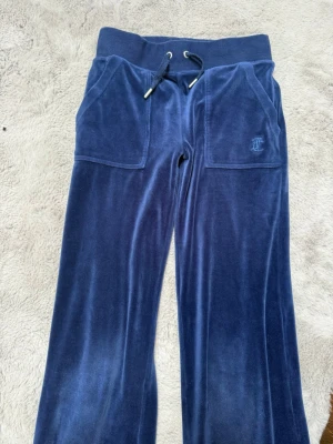 Blå velourbyxor Juicy Couture - Mörkblå mjukisbyxor i velour från Juicy Couture med bred resår i midjan och dragsko. Byxorna har stora fickor framtill och broderad JC-logga på ena fickan. Perfekta för en chill och avslappnad stil.