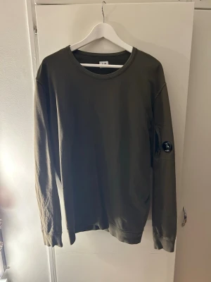 Olivgrön sweatshirt från C.P. Company - Snygg olivgrön sweatshirt från C.P. Company med rund halsringning och lång ärm. Tröjan har en ikonisk linsdetalj på ärmen och ribbade muddar vid ärmslut och nederkant. Perfekt för en avslappnad streetwear-look.