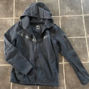 Svart Nike Tech Fleece - Snygg svart tech fleece från Nike med dragkedja framtill och huva. Den har diskreta svarta detaljer över bröstet och en praktisk ficka med dragkedja på ena ärmen. Tillverkad i ett mjukt och bekvämt material, perfekt för en avslappnad stil. Superbra kvalité, nästan helt oanvänd. Postar inom 1-2 dagar. Pris ej hugget i sten så kom med bud.