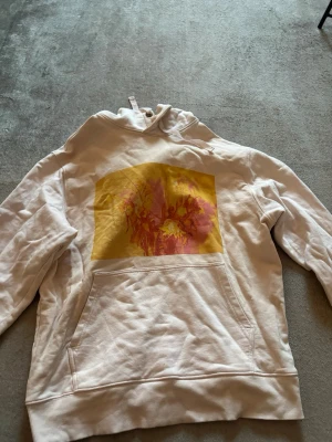 Vit hoodie med tryck från Weekday - Säljer en rosa hoodie från Weekday i storlek M. Den har en stor ficka framtill och ett färgstarkt tryck i gult och rosa på bröstet. Hoodien har huva med dragsko och är tillverkad i mjukt bomullsmaterial. Perfekt för dig som gillar streetwear och vill sticka ut.