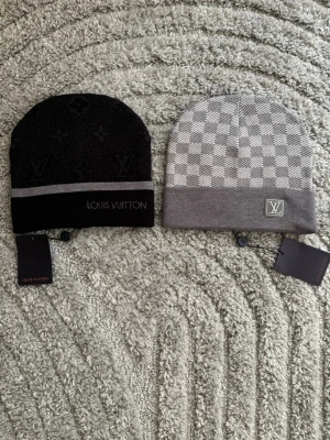 Grå och svart mössa - Två snygga mössor, en i svart med diskret monogrammönster och silvergrå rand, samt en i grått med rutigt mönster. Båda är stickade och har vikbar kant. Perfekta accessoarer för att lyfta din stil. Diskutera för att köpa en mössa enskild.