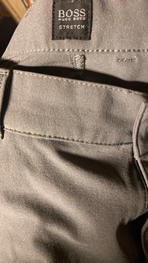 Grå stretch-chinos från Hugo Boss - Snygga grå chinos från Hugo Boss i modellen PHOENIX. Tillverkade i Italien av 92% nylon och 8% elastan för en riktigt skön stretchig känsla. Klassisk passform med bälteshällor och diskreta sömmar. Perfekta för dig som vill ha stilrena och bekväma byxor.