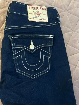 True Religion mörkblå bootcut jeans - Säljer ett par mörkblå jeans från True Religion med ikonisk kontrastsöm och stor ficka bak med lock och knapp. Modellen har bootcut-ben och klassisk femficksdesign. Perfekta för dig som gillar streetwear och vill sticka ut med detaljer.