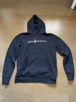Svart hoodie från Sail Racing i mycket gott skick - Svart hoodie från Sail Racing med vit logga tryckt på bröstet. Klassisk modell med huva och känguruficka framtill. Tillverkad i mjukt material som känns skönt mot huden. Perfekt för en avslappnad och sportig stil. Tröjan är knappt använd och nästan i nyskick, tröjan kosta från början 1000kr har jag för mig.