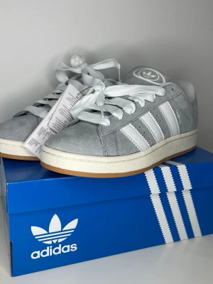  Adidas campus grå - Snygga grå sneakers från Adidas med klassiska vita ränder på sidorna och Adidas-logga på hälen. Skorna är låga och har ovandel i mjuk mocka, vita skosnören och en vit sula med gummidetalj. Perfekta för dig som gillar stilren och sportig look. Aldrig använd 