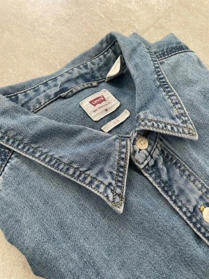 Levi's ljusblå jeansskjorta - Klassisk ljusblå jeansskjorta från Levi's med lång ärm, bröstficka och knappar framtill. Skjortan har en avslappnad passform och är tillverkad i mjuk bomull. Perfekt för en cool och tidlös look.