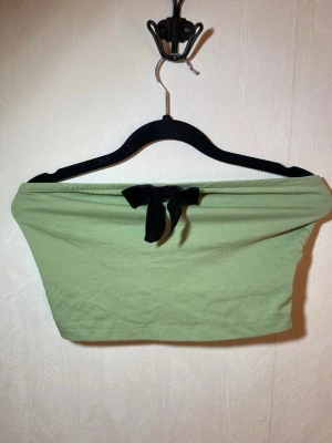 Grön axelbandslös tubtopp med rosett - Snygg ljusgrön axelbandslös tubtopp med en svart sammetsrosett framtill. Perfekt för dig som vill ha en enkel men ändå söt topp till sommaren. Mjuk och stretchig känsla. Helt ny, oanvänd. Shein, storlek M. Djur, rök och parfymfritt hem.