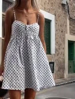 Vit prickig sommarklänning - Söt vit kortklänning med svarta prickar och tunna axelband. Klänningen har en lätt utställd passform och markerad midja, perfekt för varma dagar. Tillverkad i ett mjukt och luftigt material som känns skönt mot huden.