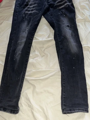 Svarta distressed jeans från Dsquared2 - Säljer ett par svarta jeans från Dsquared2 i storlek 52. Jeansen har en slim fit, slitningar och färgstänk för en riktigt edgy look. Tillverkade i bomull med klassisk femficksmodell och knappgylf. Perfekta för dig som gillar streetwear och trendiga detaljer. Passar perfekt på mig och är 183cm och 65kg