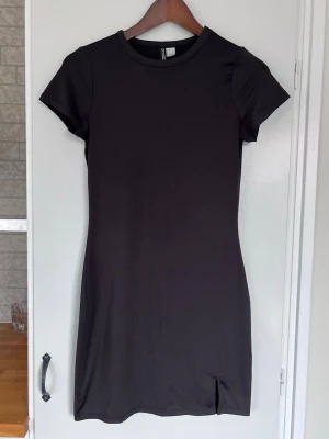 Svart miniklänning från H&M, stl S - En kort, tight t-shirtklänning från H&M i tunt skönt och stretchig material. En liten slits nertill på vänster lår. Sparsamt använd, mycket bra skick!