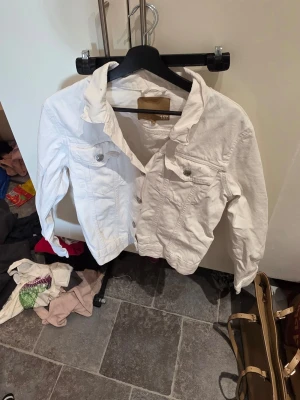 Vit jeansjacka från 157 - Snygg vit jeansjacka från 157 med klassisk krage, två bröstfickor med knappar och silverfärgade knappar framtill. Jackan har en rak passform och långa ärmar. Perfekt att slänga över en t-shirt för en cool och avslappnad look.