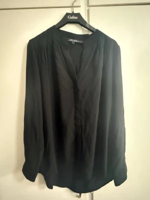 Svart blus med v-ringning från Marc Lauge - Stilren svart blus med v-ringad hals och avslappnad passform. Blusen har långa ärmar och är tillverkad i ett lätt, mjukt tyg som ger ett snyggt fall. Perfekt att matcha med jeans eller kjol för en trendig look.