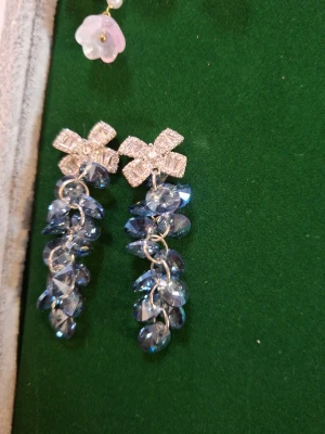 Glittriga örhängen med blå pärlor - Säljer ett par statement-örhängen med gnistrande blå pärlor och dekorativa rosetter i silverfärg upptill. Örhängena har en droppformad design med flera pärlor som hänger i kedjor. Perfekta för dig som vill sticka ut med accessoarer.