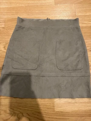 Grå mockaliknande minikjol - Snygg grå minikjol i mockaliknande material med två stora fickor framtill. Kjolen har en rak passform och dragkedja bak. Perfekt att matcha med en stickad tröja eller t-shirt för en trendig look.