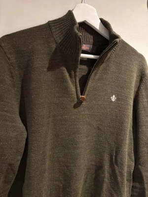 Olivgrön stickad tröja med halvzip - Snygg olivgrön stickad tröja från Morris med halvzip och hög krage. Tröjan har ribbade muddar och en liten vit logga broderad på bröstet. Perfekt för lager på lager och en stilren look.