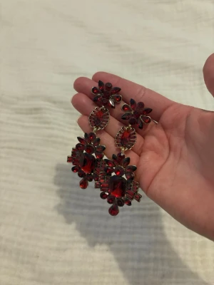 Stora röda statement örhängen - Säljer ett par stora statement örhängen med gnistrande röda stenar i olika former. Örhängena har en antikguldig metallbas och är dekorerade med både runda och avlånga stenar som ger en lyxig och iögonfallande look. Perfekta för dig som vill sticka ut och addera färg till din outfit.