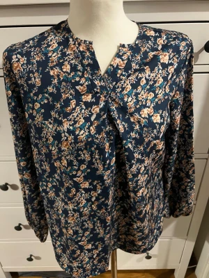 Mörkblå blommig blus med V-ringning - Snygg mörkblå blus med småblommigt mönster i beige, vitt och turkost. Blusen har V-ringning med dekorativ vikt detalj framtill och långa ärmar. Tillverkad i ett lätt och mjukt tyg som ger en skön känsla.