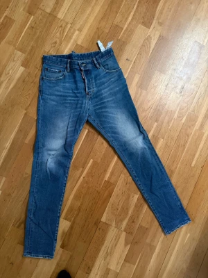Blå jeans från Dsquared2. Storlek 48. - Säljer ett par blå jeans från Dsquared2 med klassisk femficksmodell och snygga slitningar på låren. Jeansen har smal passform och är tillverkade i denim med tydliga kontrastsömmar. Perfekta för en trendig och avslappnad stil. Finns i Helsingborg. Nypris var cirka 4100kr. 