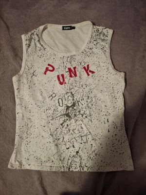 Vit ärmlös punk rock linnetopp - Cool vit linnetopp från Cami med svart splattermönster och texten 'PUNK ROCK' i rött och svart framtill. Toppen är ärmlös och har en rund halsringning. Perfekt för dig som gillar en edgy och avslappnad stil.