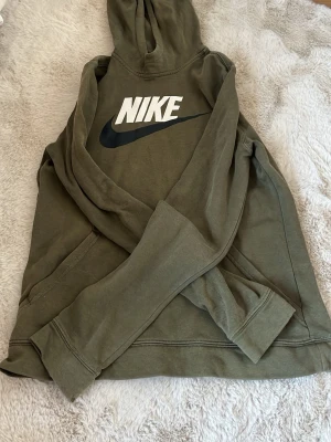 Olivgrön Nike hoodie med logga - Säljer en olivgrön hoodie från Nike med stor vit och svart logga på bröstet. Hoodien har huva och ribbade muddar vid ärmslut och nederkant. Tillverkad i mjukt bomullsmaterial, perfekt för en avslappnad streetwear-look.