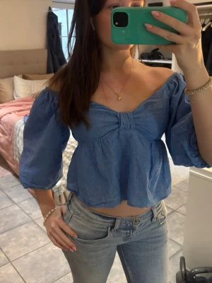 Blå offshoulder blus med puffärm - Trendig blå blus med offshoulder och trekvarts puffärmar. Croppad modell med lätt rynk och söt detalj framtill. Perfekt till jeans eller kjol för en cool look. Troligtvis i bomull eller linne för en luftig känsla.