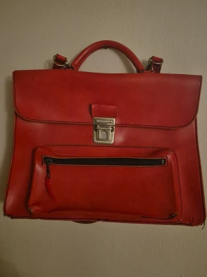 Röd portfölj i läder med axelrem - Snygg röd vintage portfölj i läder med både handtag och avtagbar axelrem. Väskan har ett stort huvudfack, en frontficka med dragkedja och ett metallspänne framtill. Perfekt för laptop, böcker eller viktiga papper. Klassisk och stilren design. Strl 31 x 24 cm. Är generellt i renoveringsbehov med sömmar som brustit i hörnen. Men en snygg retro/vintage väska för den som orkar. Finns inget varumärke 