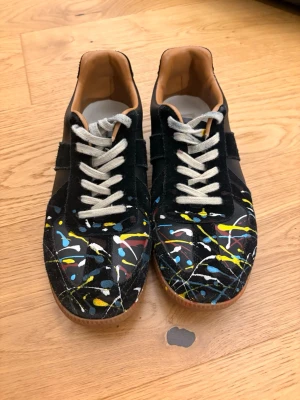 Svarta sneakers med färgstänk - Unika svarta sneakers från Maison Margiela med coola färgstänk i gult, blått, rött och vitt. Skorna har vita snören, bruna innersulor och gummisula. Materialet är en mix av mocka och läder för en lyxig känsla och streetstyle-vibe.